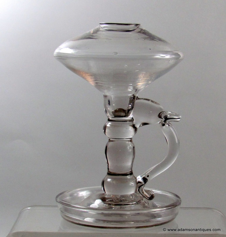 Clear glass, oil-burning chamber lamp with a handle on one side and a saucer beneath the bowl and stem. 